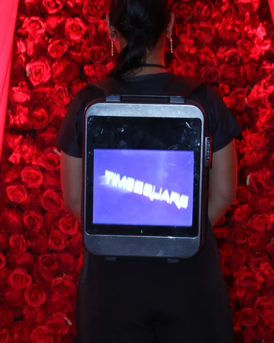 An LCD backpack worn from behind against a wall of red roses, its screen showing the Timesquare logo