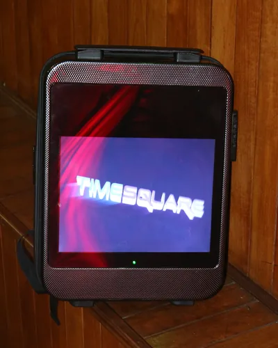 An LCD backpack standing on a wooden bench, its screen showing the Timesquare logo