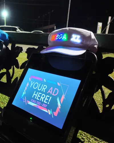 Two LCD backpacks on a park bench at night reading Your Ad Here, with a white LED cap on top lit with scrolling lettering