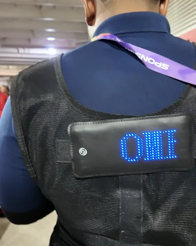 The back of a crew member's black LED vest at the CPL 2025 ground, its blue pixel panel scrolling the sponsor name