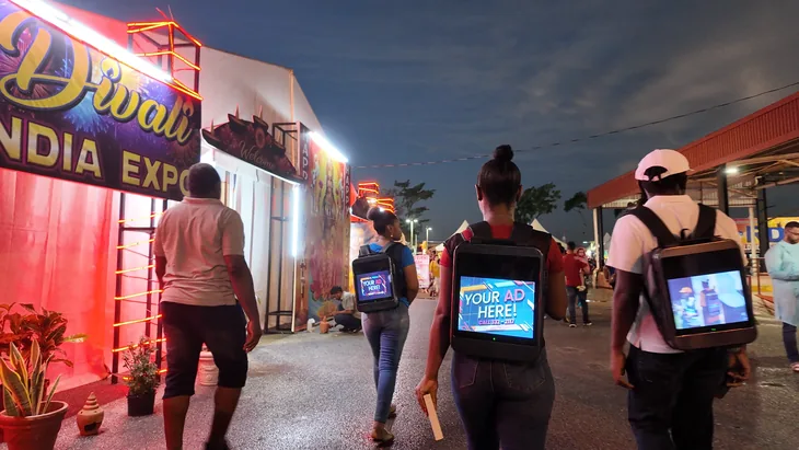 Two people walking away through the Divali Nagar festival grounds at dusk past the Divali India Expo sign, each wearing a lit Timesquare LCD backpack reading Your Ad Here