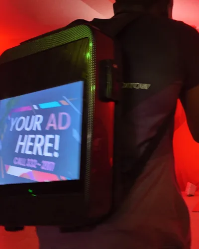 An LCD backpack reading Your Ad Here worn in a ballroom washed in red light, a rose-wall arch behind